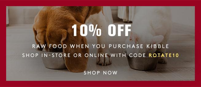 A basset hound and a white cat eating out of metal bowls. 10% off raw food when you purchase kibble. Shop in-store or online with code: ROTATE10. Shop Now