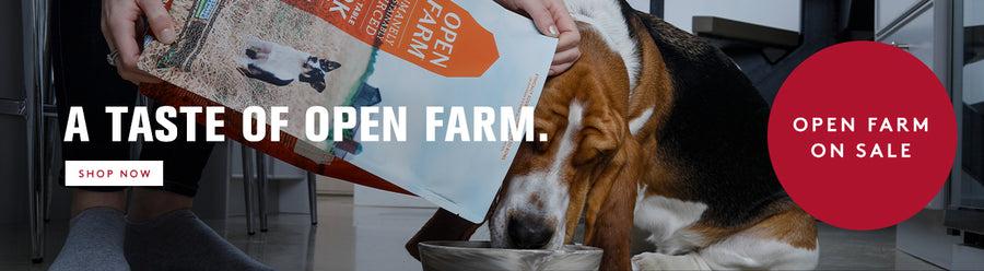 A basset hound eating open farm dog food out of a silver bowl while the owner is pouring in open farm food from the bag. A TASTE OF OPEN FARM.  enjoy Open Farm deals all month long